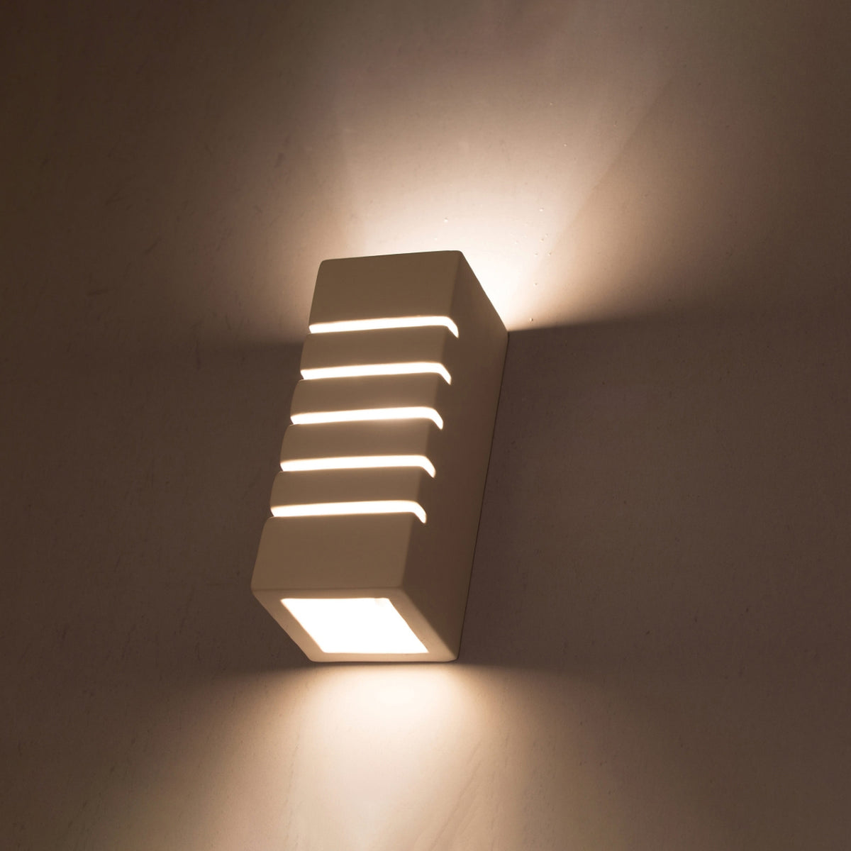 Applique allumée sur un mur, lumière chaude filtrant par les fentes horizontales.