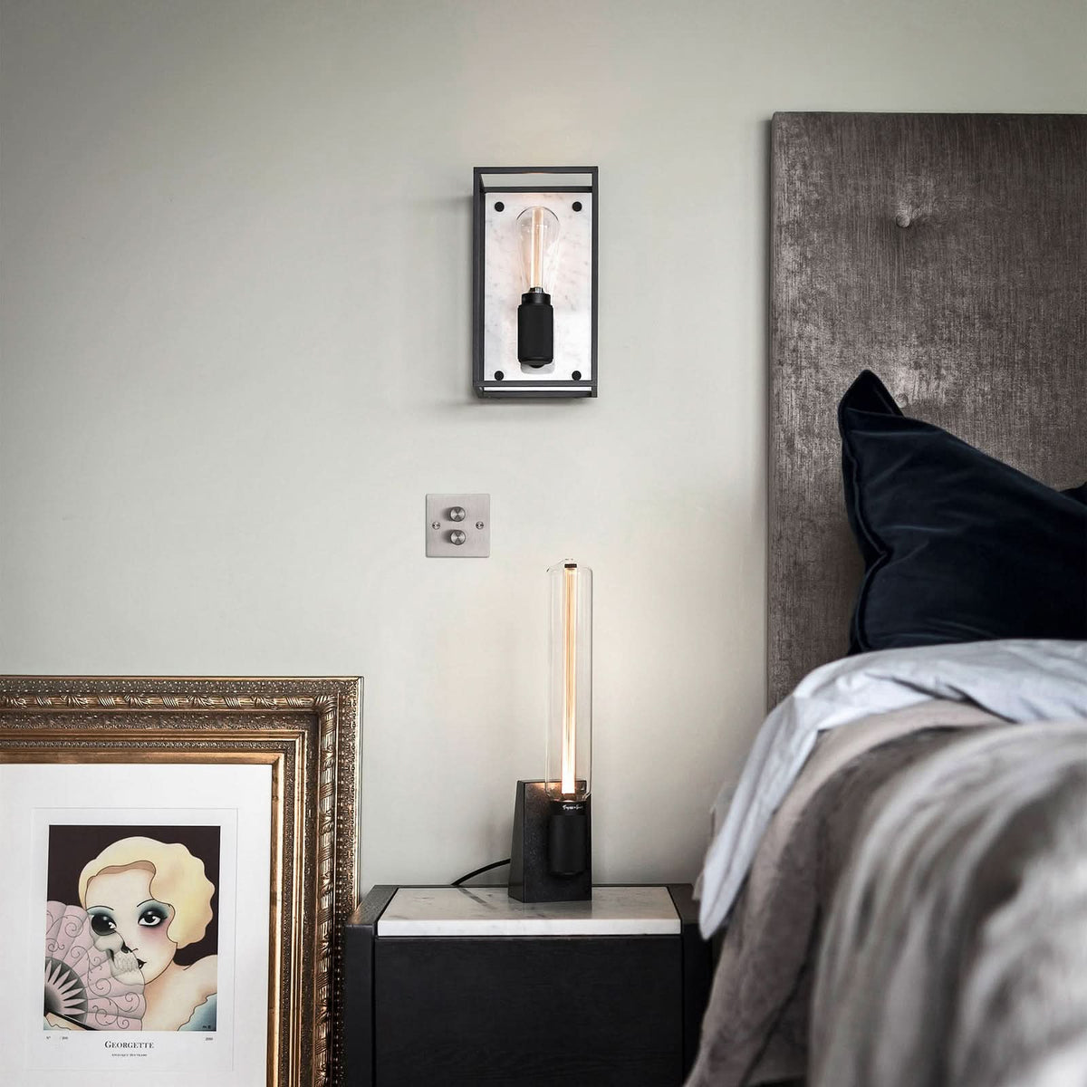 Mise en ambiance dans une chambre : applique au-dessus d'une table de chevet, cadre et lit.