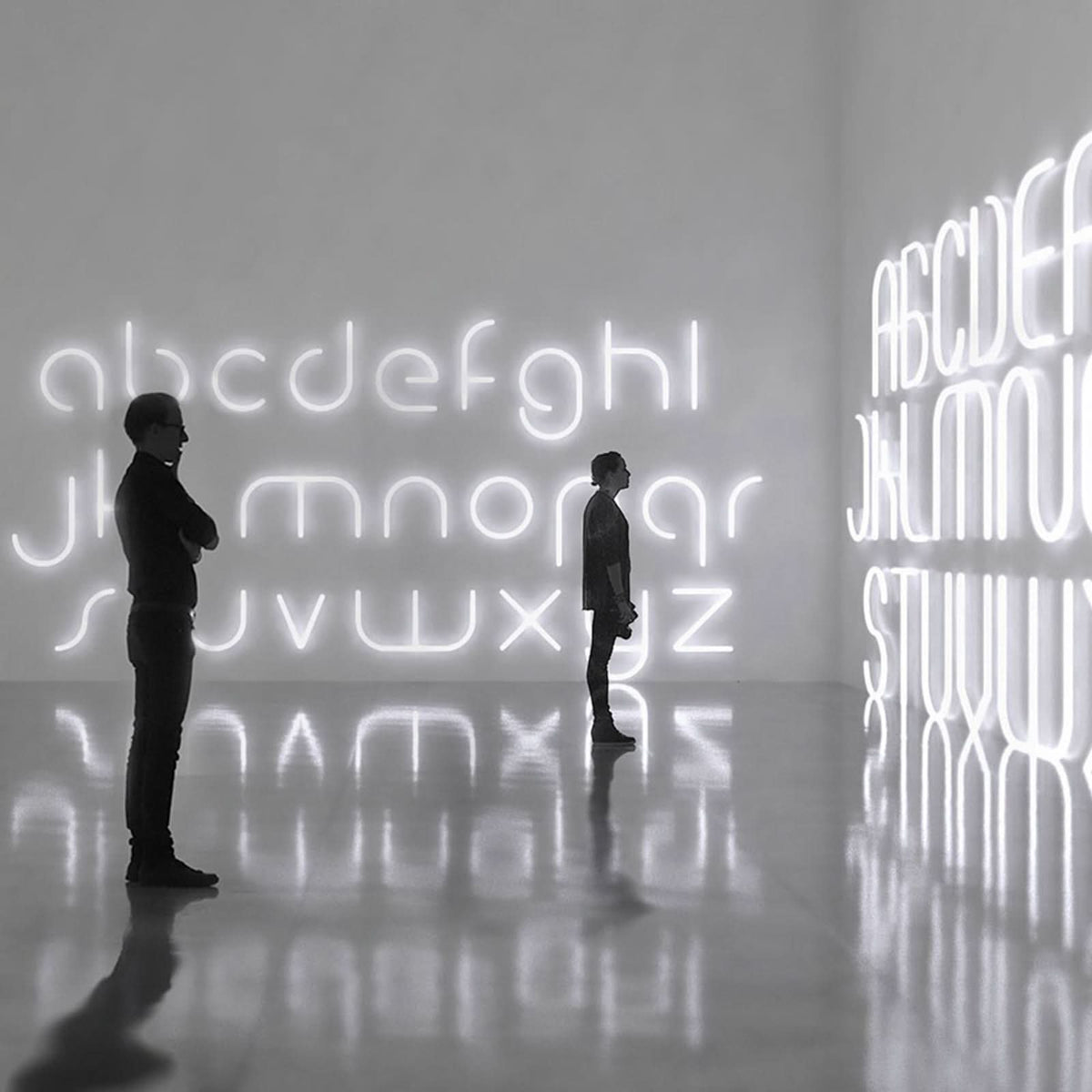 Deux silhouettes observent des lettres lumineuses blanches dans une galerie au sol réfléchissant