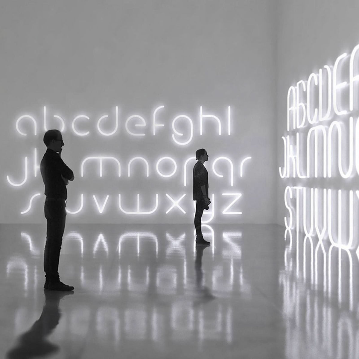 Deux personnes contemplant un mur couvert d'un immense alphabet lumineux, reflets sur le sol
