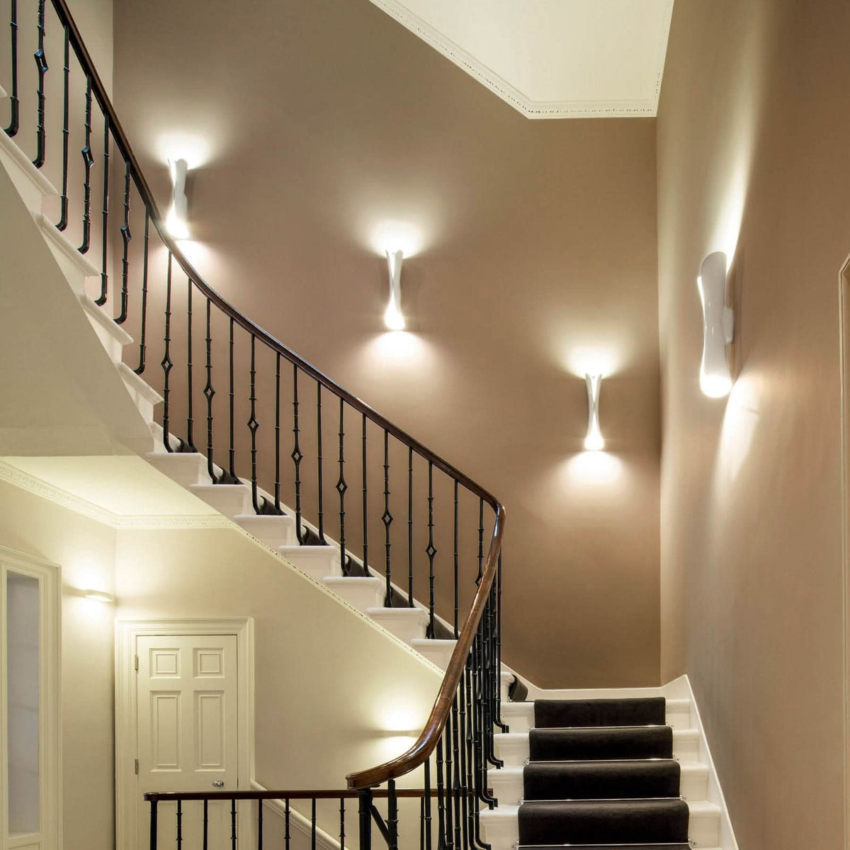 Plusieurs appliques blanches éclairent une cage d'escalier beige avec rampe en fer.