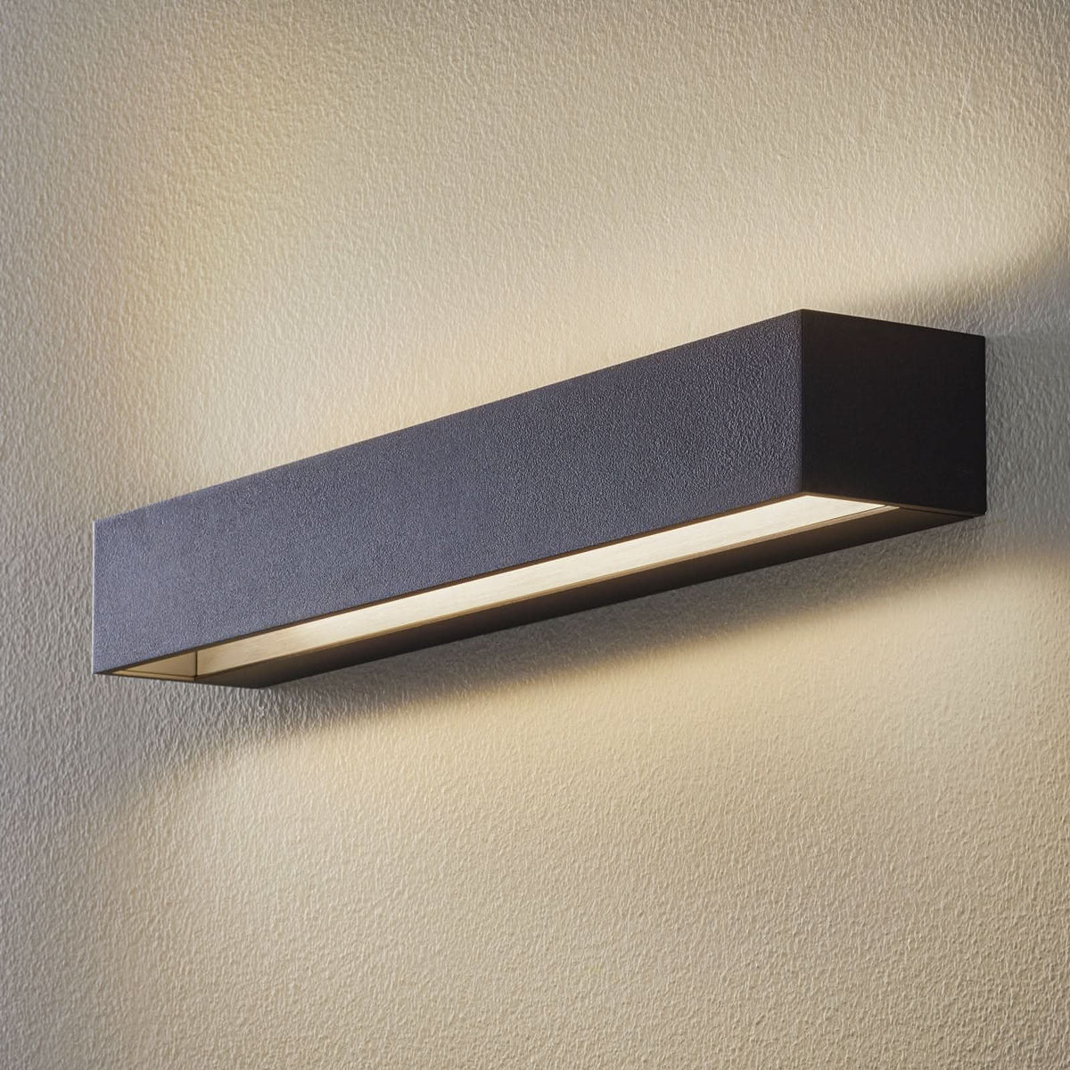 Applique murale noire rectangulaire allumée sur un mur beige texturé, lumière vers le bas.