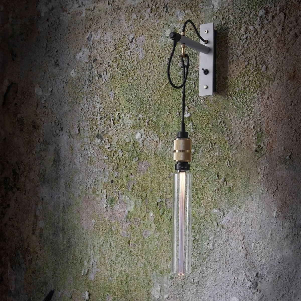 Applique murale industrielle fixée sur un mur brut, ampoule cylindrique allumée suspendue par un câble noir.