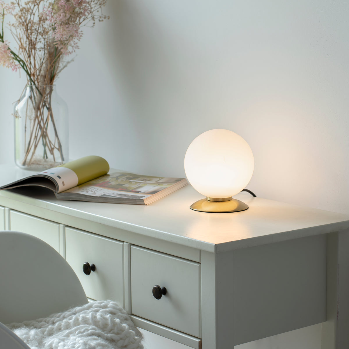 Mise en ambiance sur une commode blanche, lampe allumée près d'une revue et d'un vase.