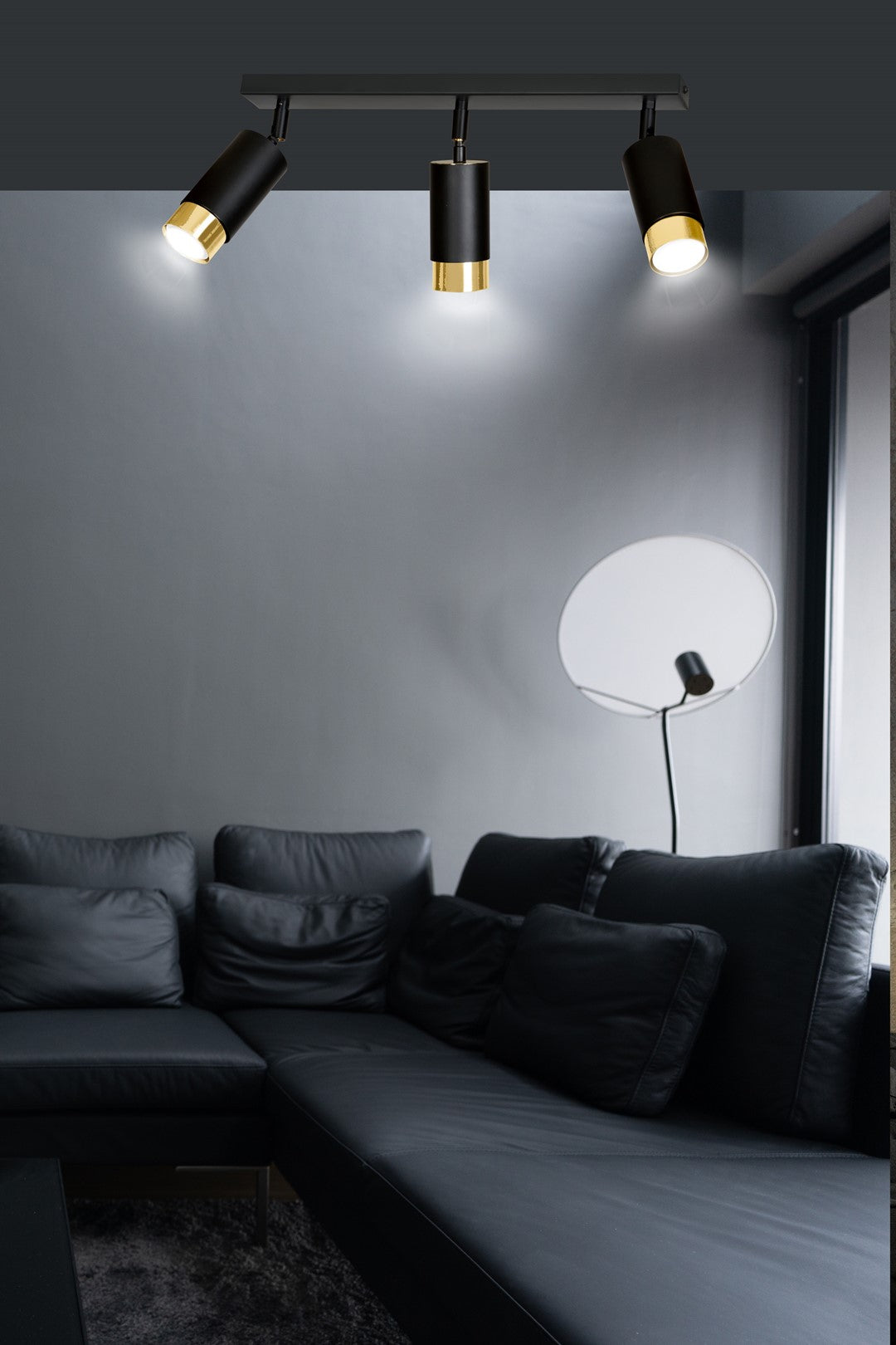 Mise en ambiance dans un salon sombre avec canapé noir et lampadaire disque blanc.