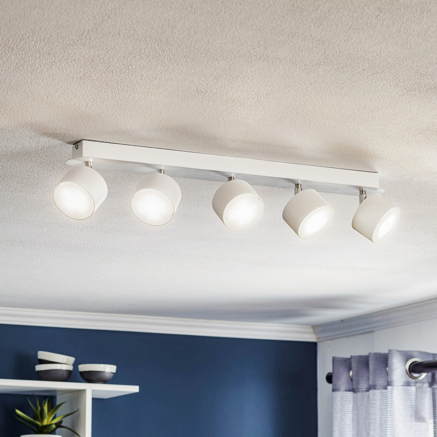 Luminaire blanc à cinq spots cylindriques fixé au plafond, allumé dans une pièce bleue.