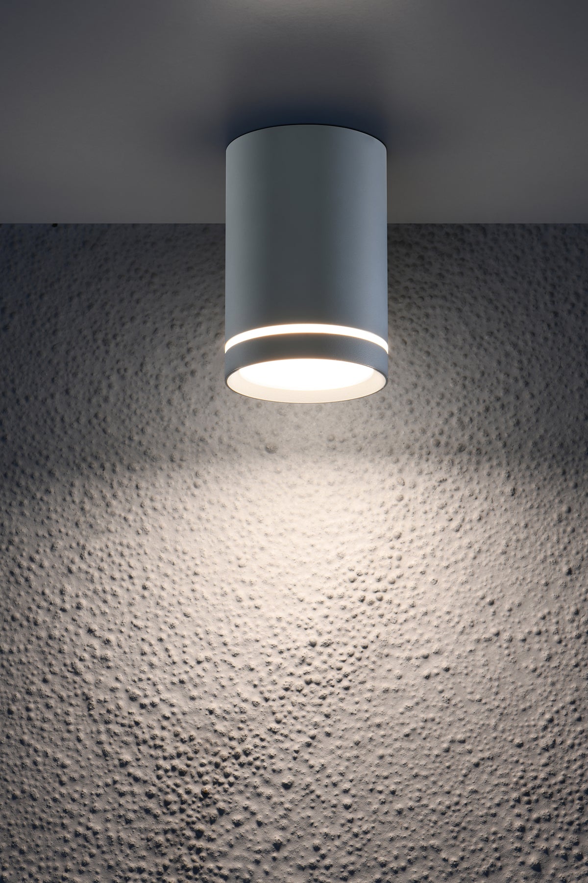 Détail en contre-plongée du luminaire gris allumé éclairant un mur texturé
