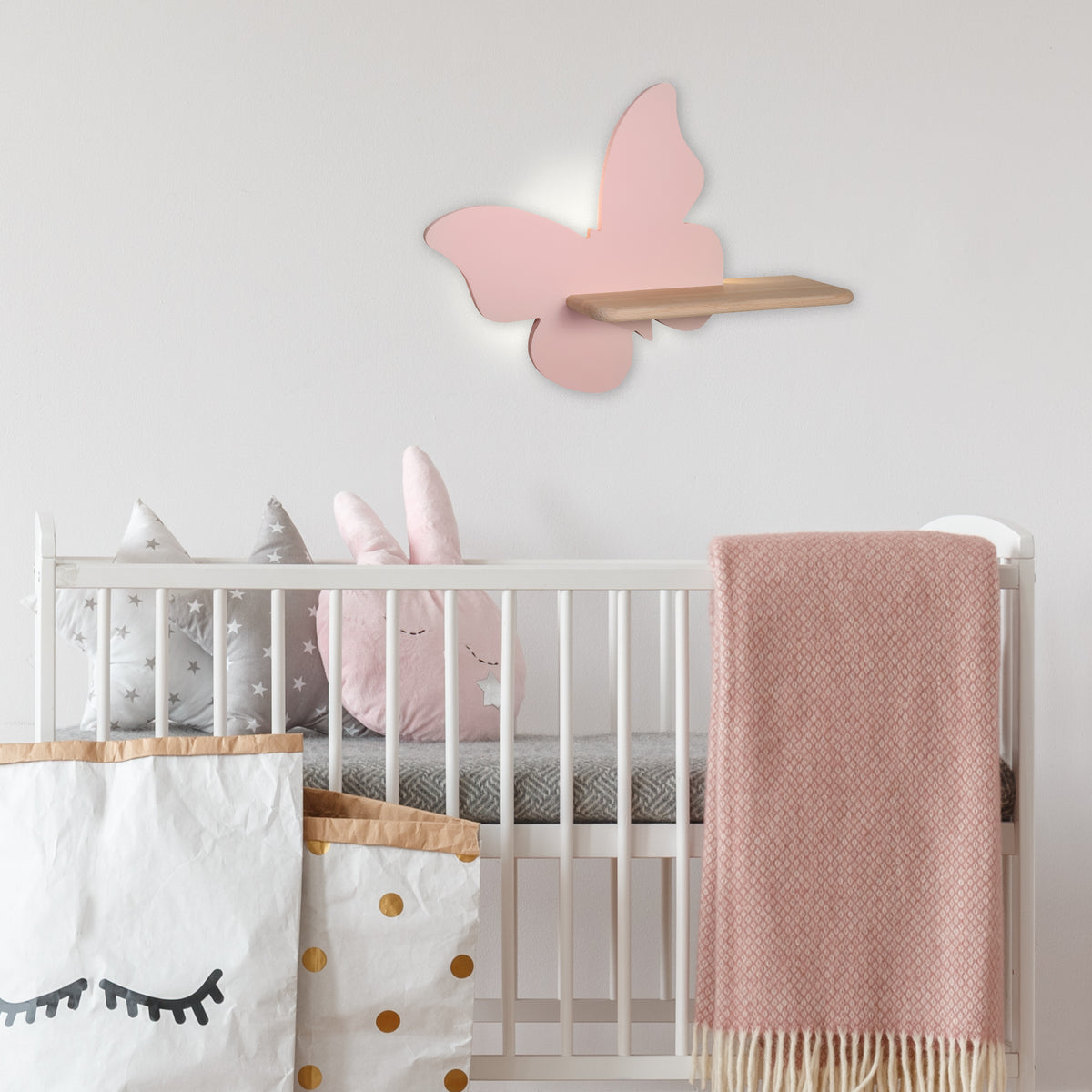 Mise en ambiance dans une chambre d'enfant : applique papillon rose allumée au-dessus d'un lit à barreaux.