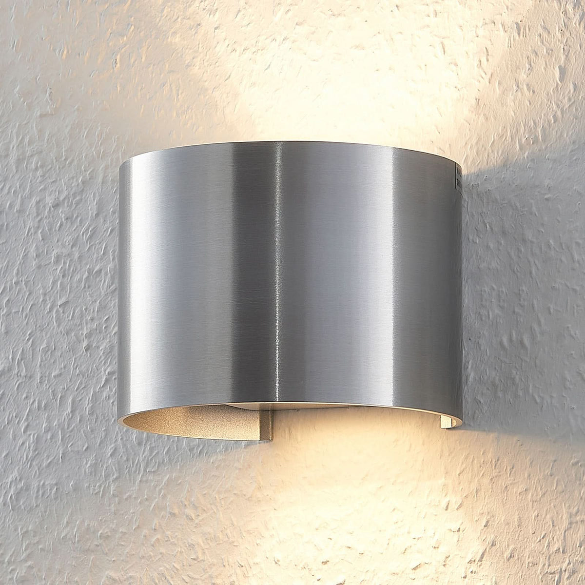 Applique murale cylindrique en aluminium brossé, montée sur un mur texturé et éclairée.