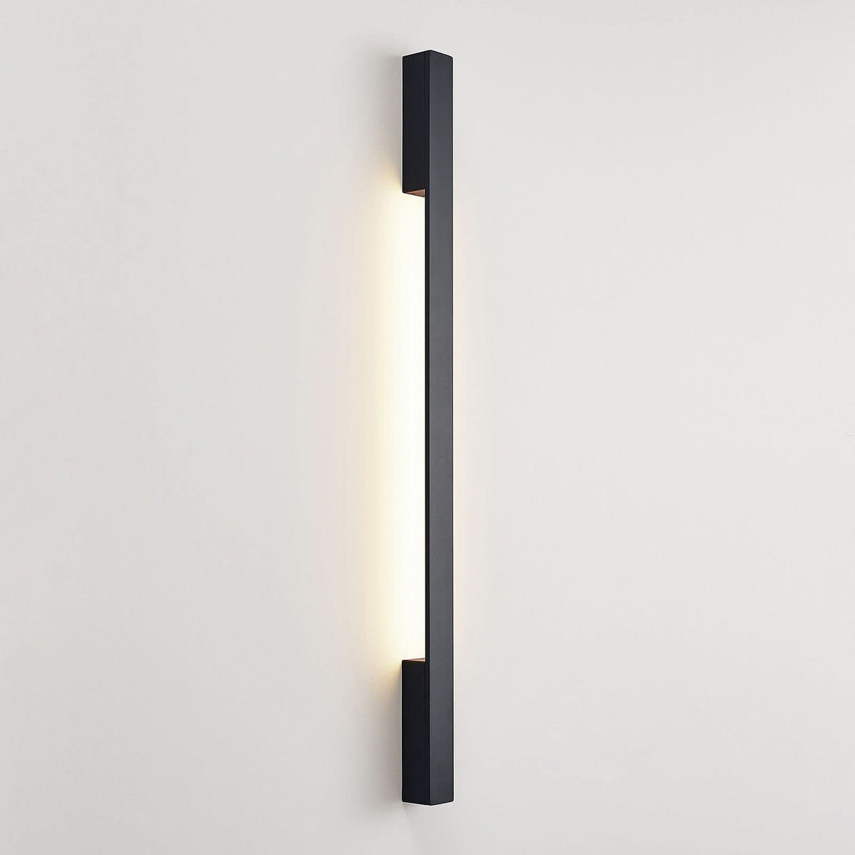Vue de face de l'applique murale LED noire allumée, halo lumineux vertical sur mur blanc.