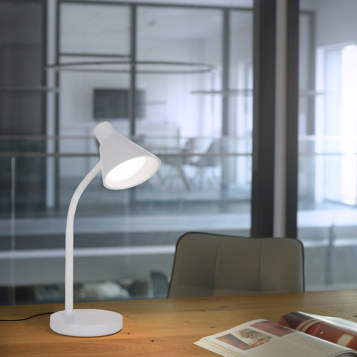 Lampe blanche allumée posée sur un bureau en bois devant une baie vitrée, ambiance bureau moderne.