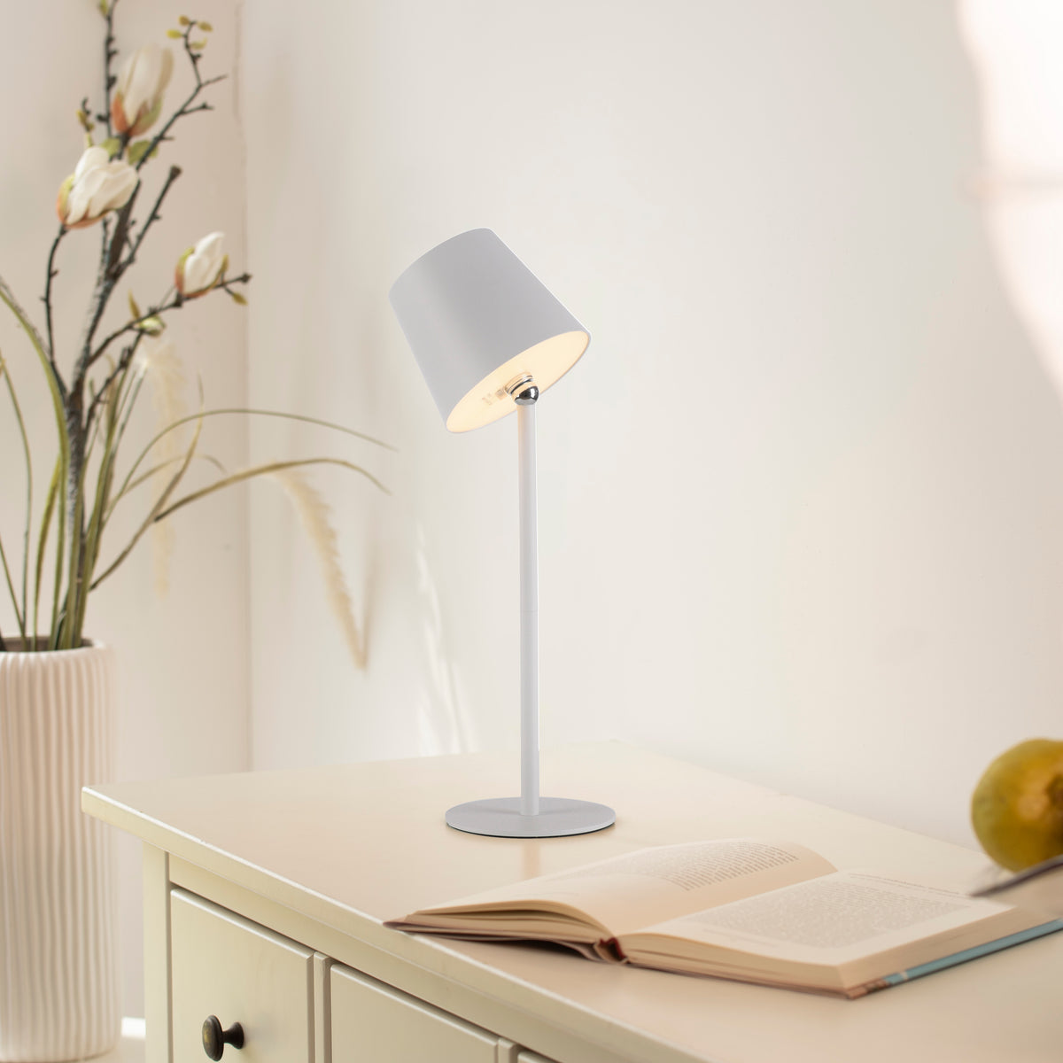 Mise en ambiance de la lampe blanche allumée sur un bureau avec livre et plante.