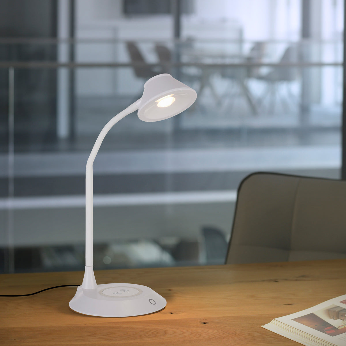 Lampe blanche posée sur une table en bois devant une salle de réunion vitrée.
