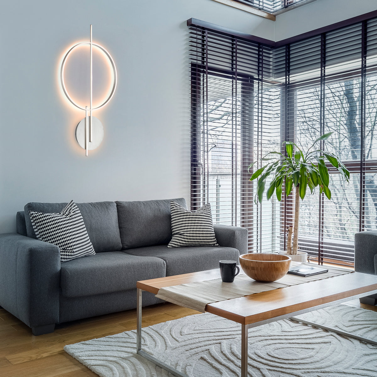Luminaire mural allumé dans un salon, au-dessus d'un canapé gris et près d'une fenêtre.