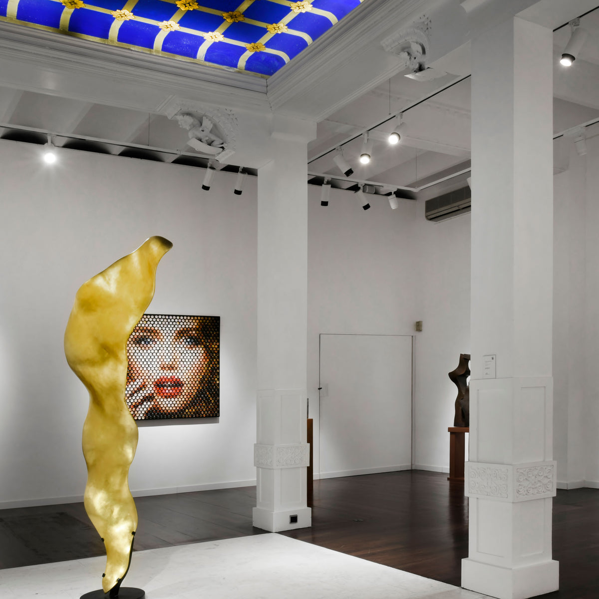 Mise en ambiance dans une galerie : plusieurs spots blancs sur rail éclairent sculptures et tableaux.