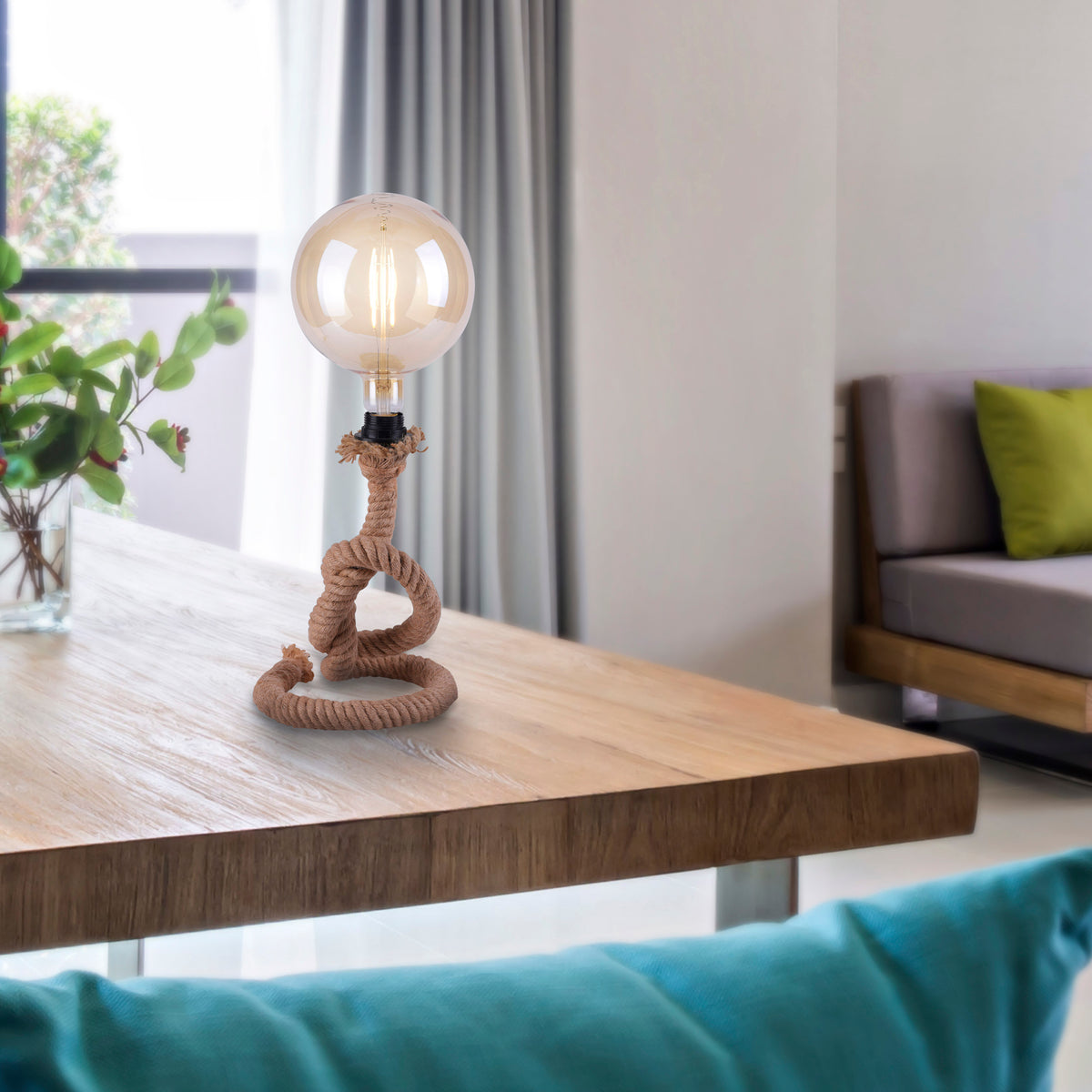 Mise en ambiance sur table en bois, lampe en corde devant canapé et plante verte