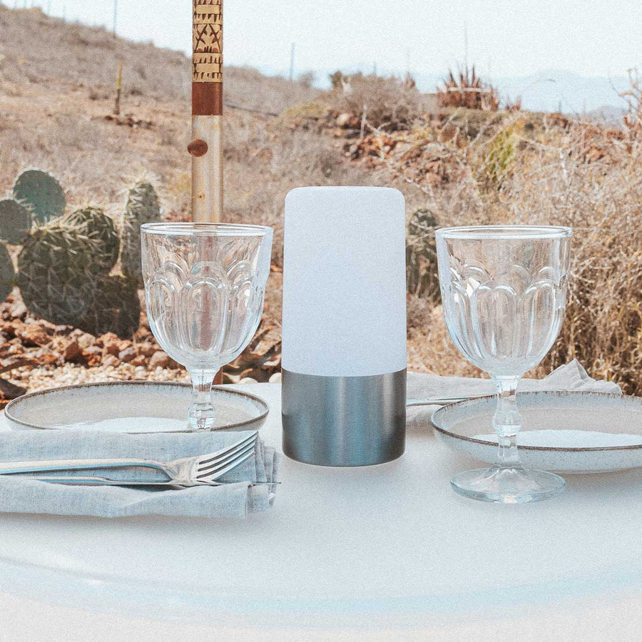 Lampe posée sur table extérieure, verres et cactus en arrière-plan désertique.