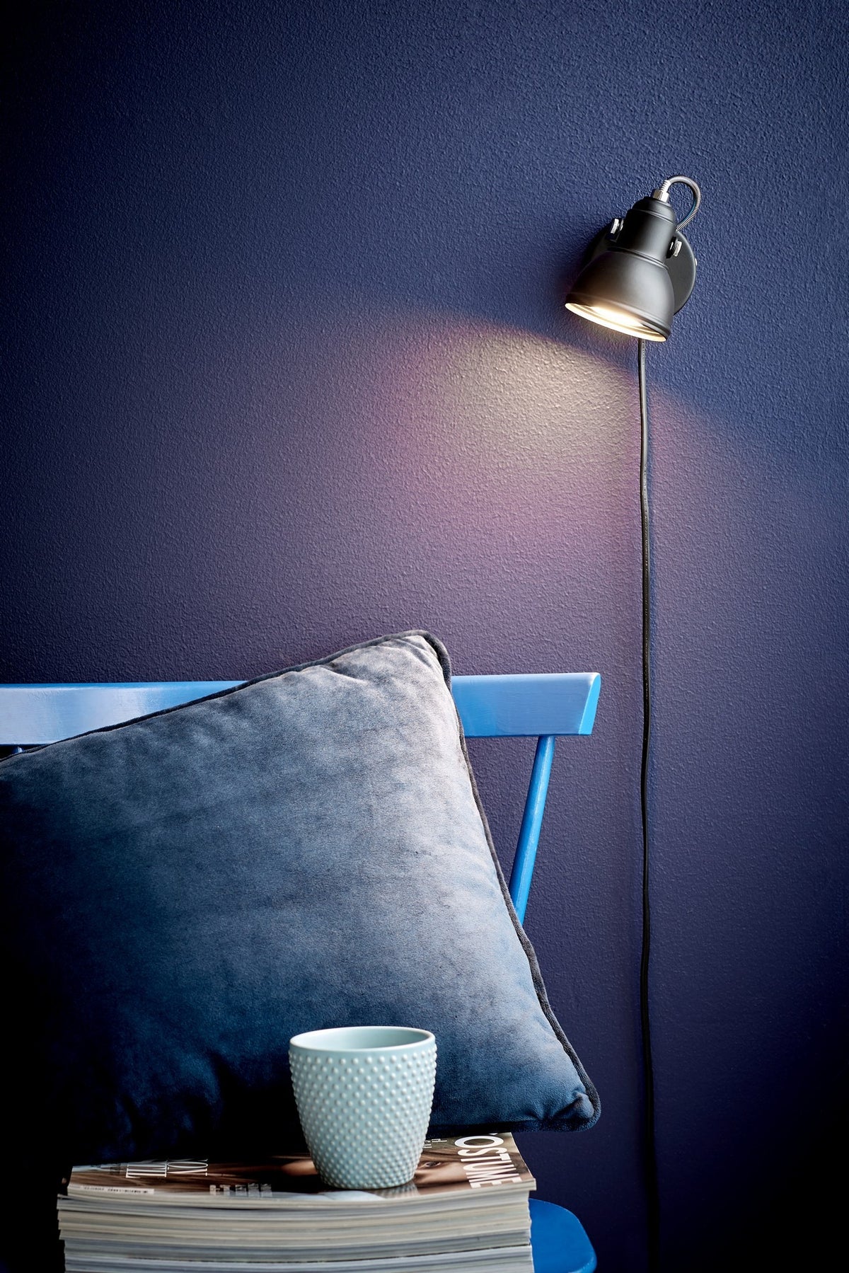 Applique noire allumée sur mur bleu, éclairant la paroi, avec cordon apparent.