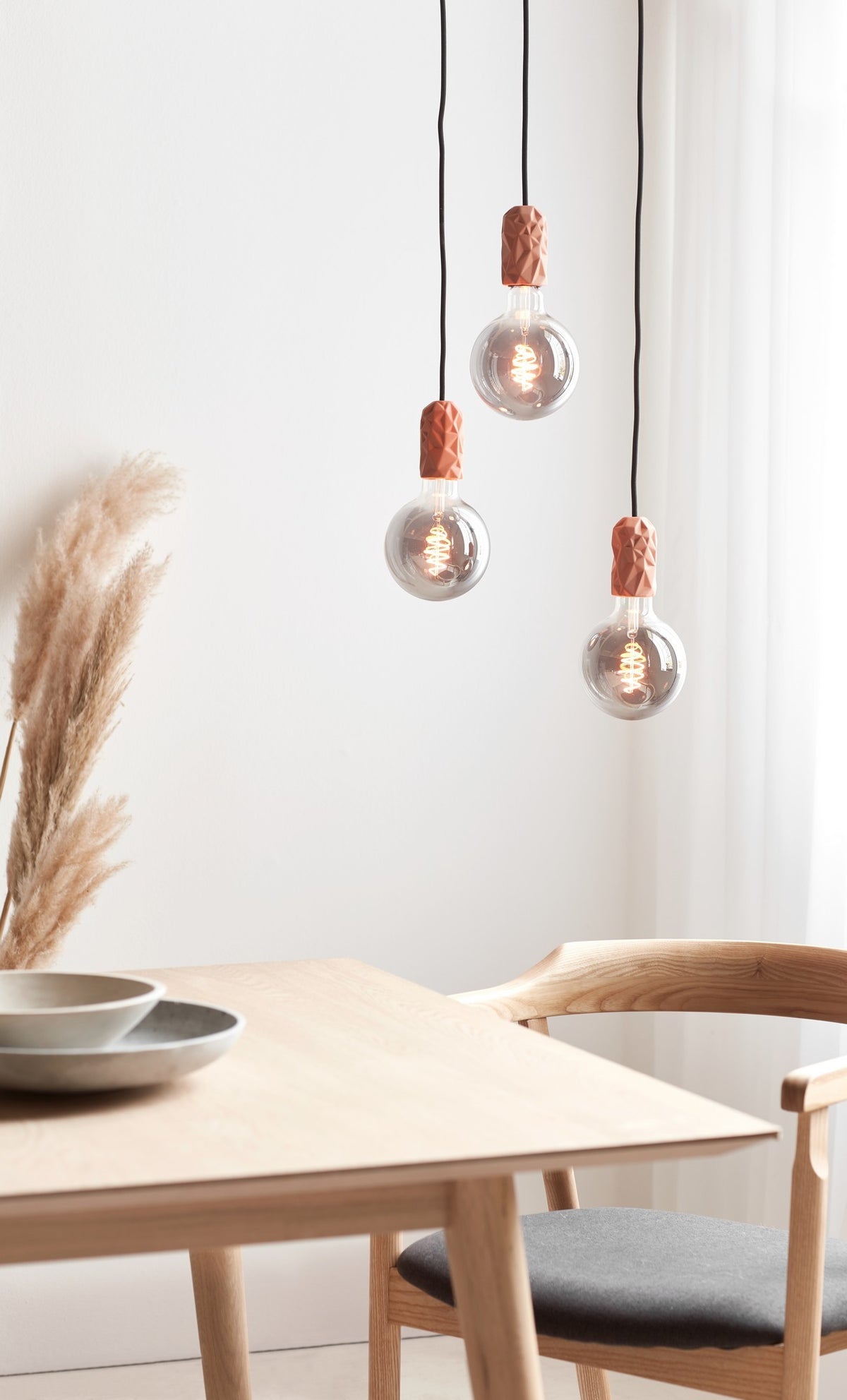 Mise en ambiance : trois suspensions terracotta au-dessus d'une table en bois avec pampas et chaise.