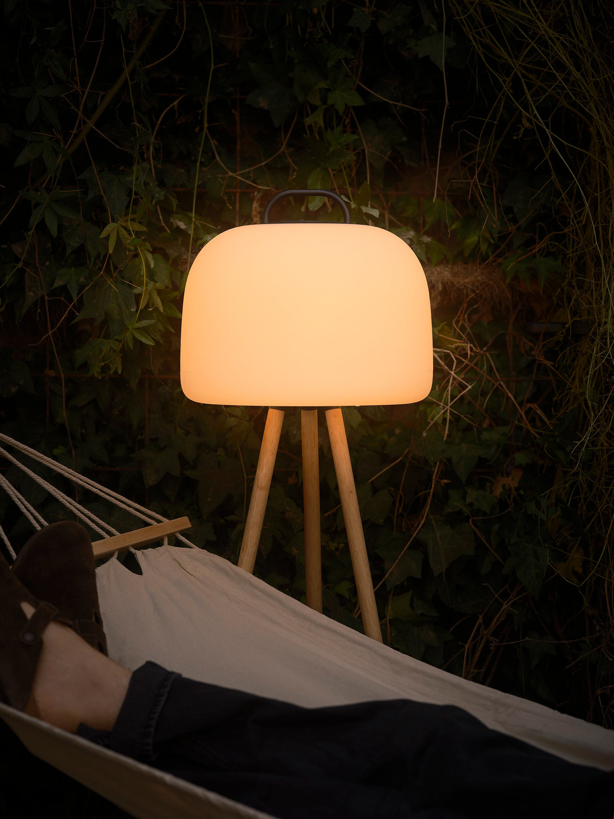 Scène de jardin, lampe allumée posée sur un trépied en bois devant des feuillages