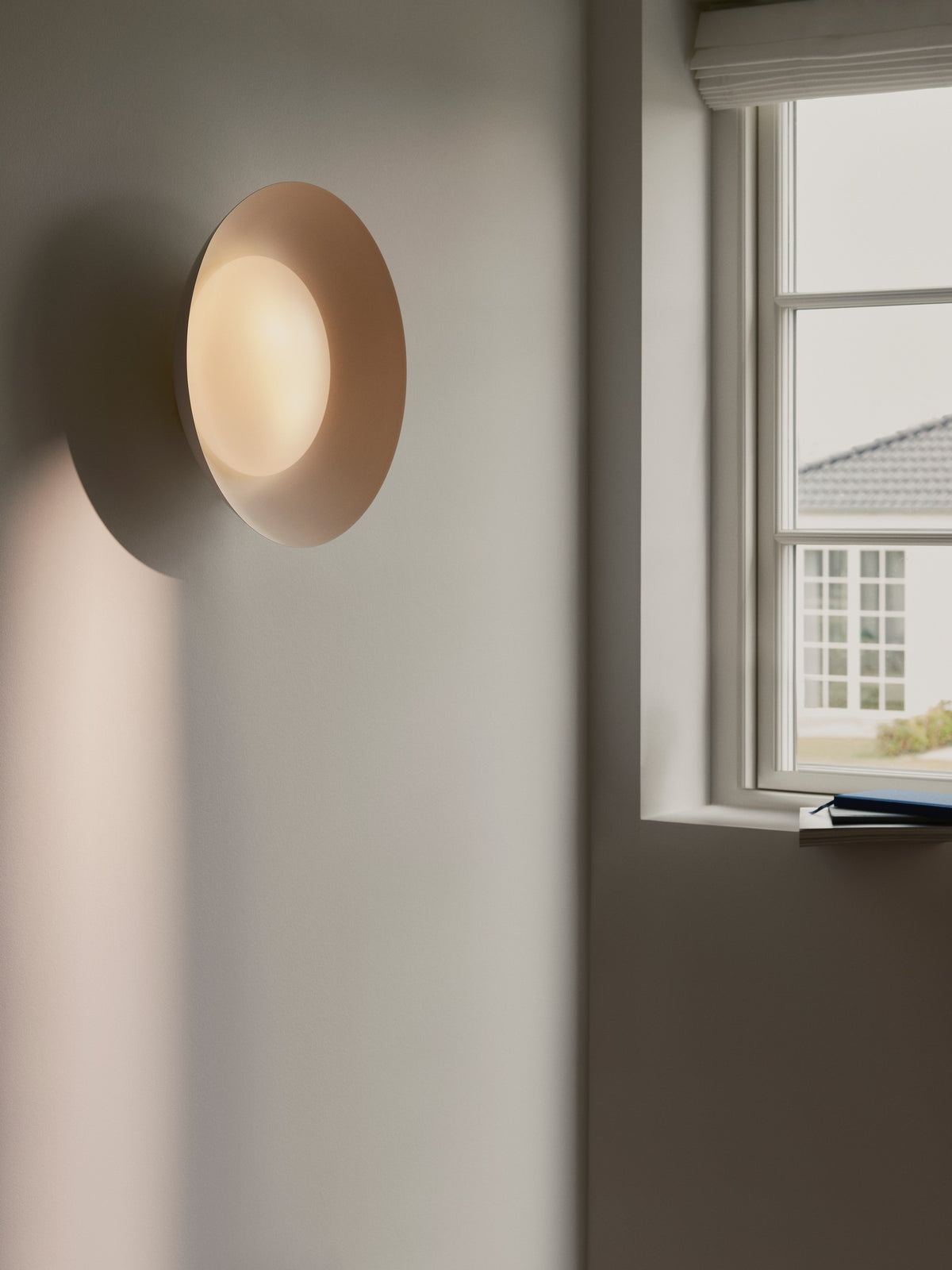 Luminaire rond blanc installé au mur près d'une fenêtre, lumière chaude allumée.