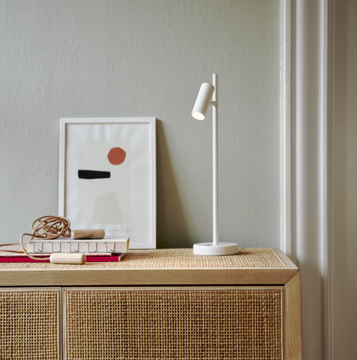 Mise en ambiance : lampe blanche posée sur un meuble en cannage, cadre et livres.