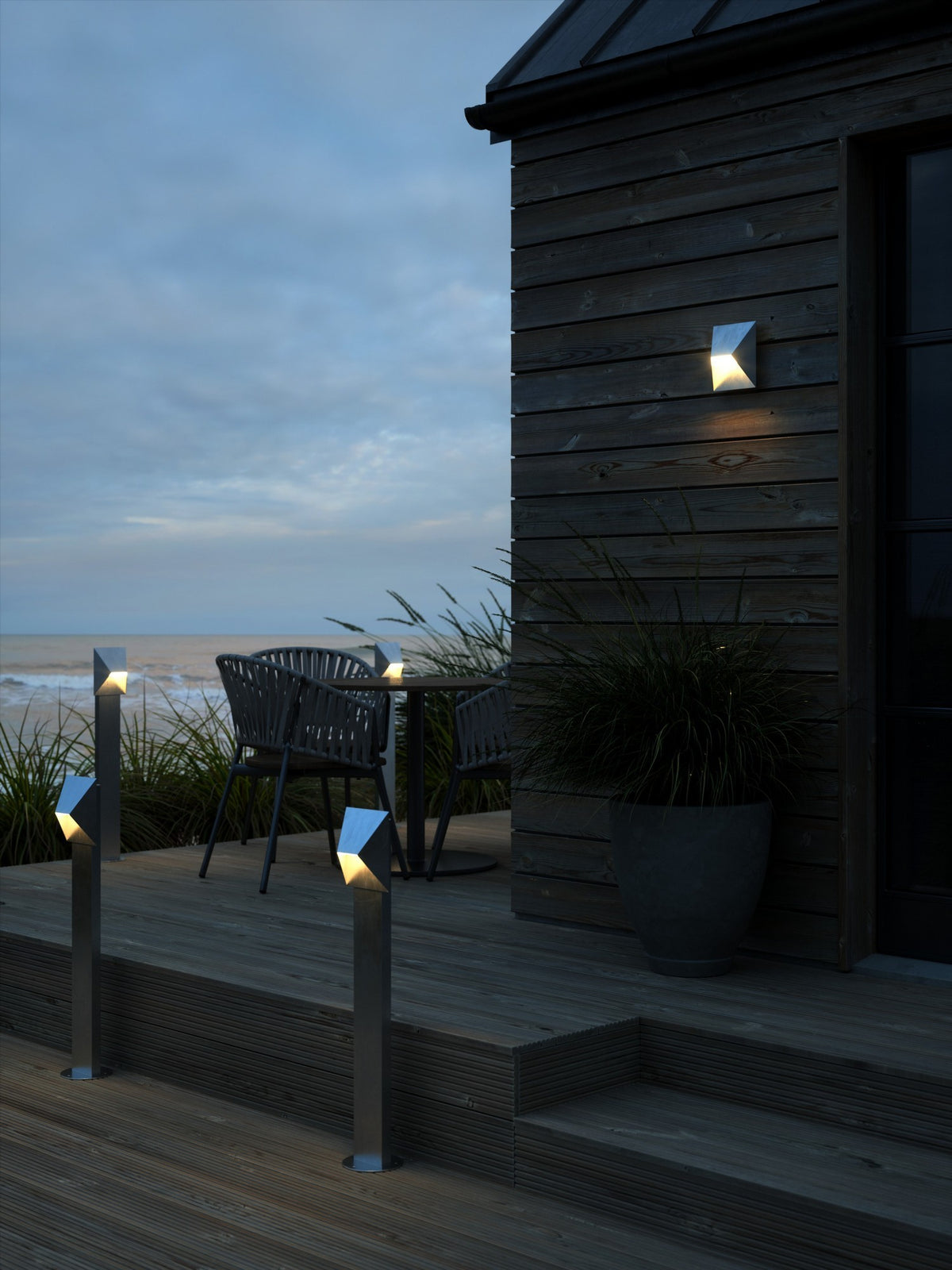 Mise en ambiance nocturne : applique et bornes éclairant une terrasse en bois face à la mer.