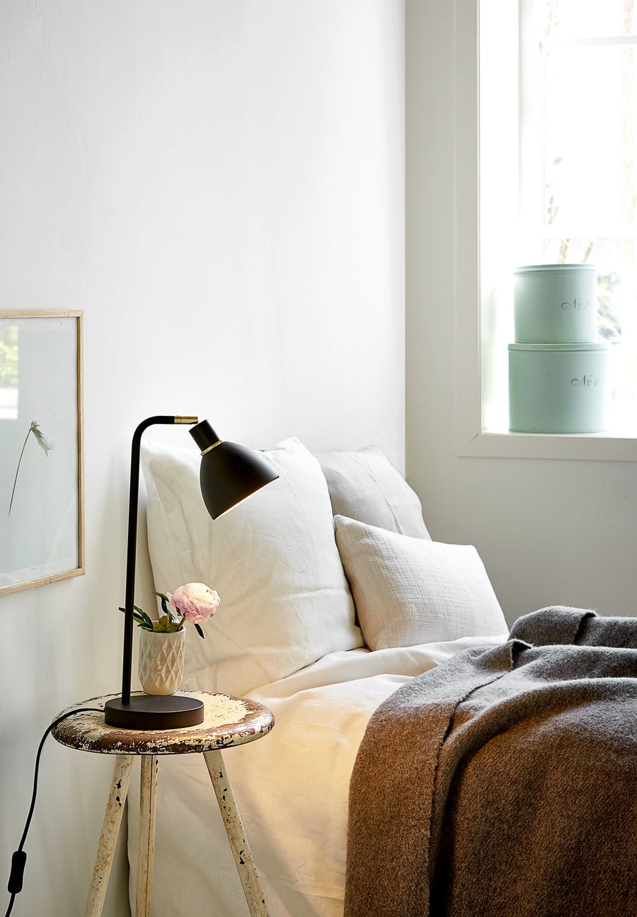 Ambiance chambre, lampe noire posée sur tabouret de nuit près du lit.