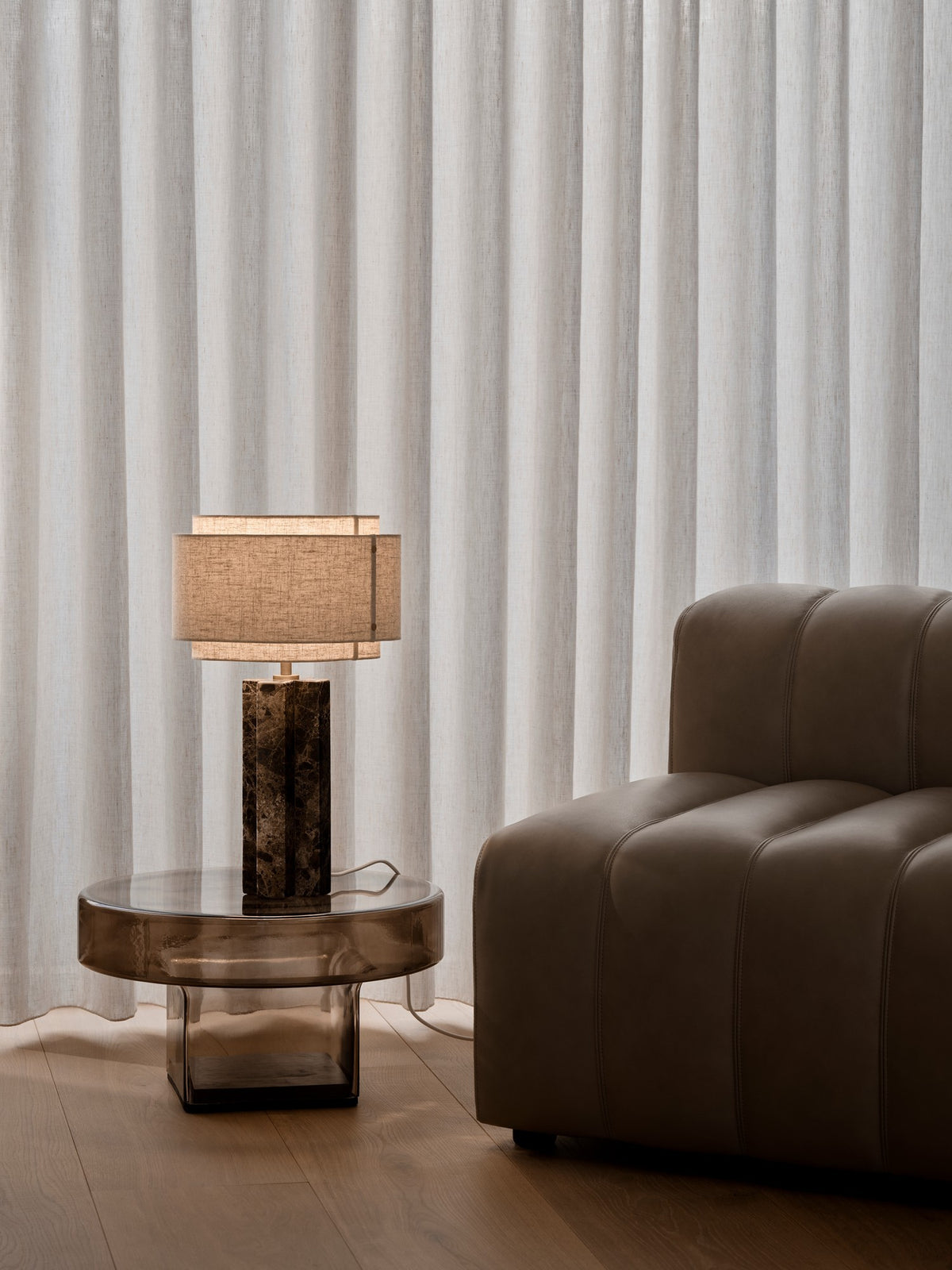 Ambiance: lampe posée sur table basse en verre, près d'un fauteuil en cuir et rideaux clairs.