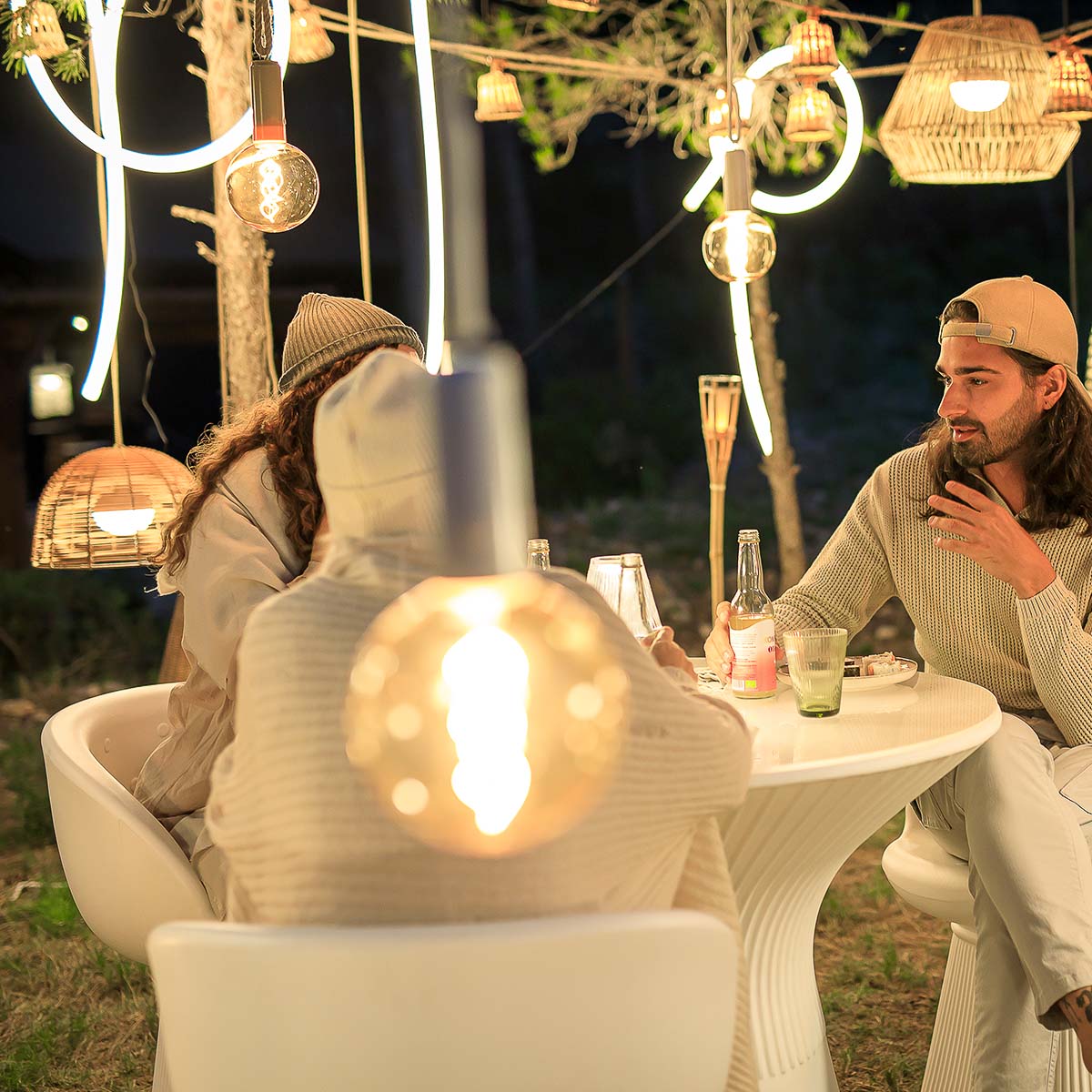 Scène conviviale en extérieur, tube LED blanc suspendu au-dessus d'une table et luminaires en rotin.
