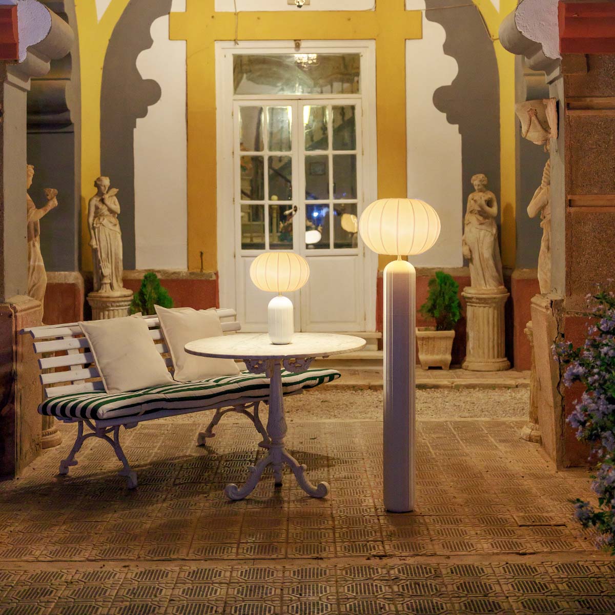 Lampadaire blanc allumé dans une cour de nuit, près d'une table et de statues.
