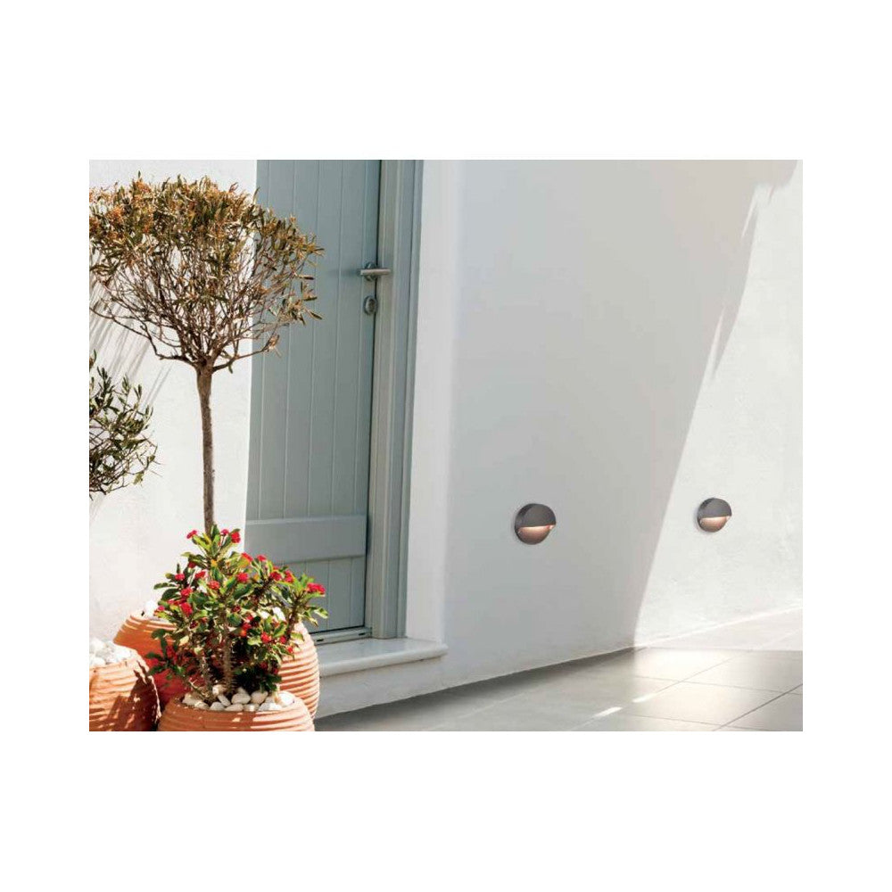 Deux appliques rondes graphite sur un mur extérieur blanc, près d’une porte et de plantes.