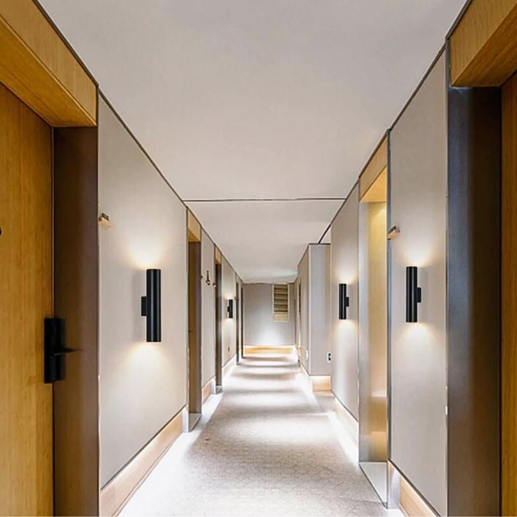 Couloir d'hôtel avec appliques noires cylindriques allumées le long des murs et portes boisées.