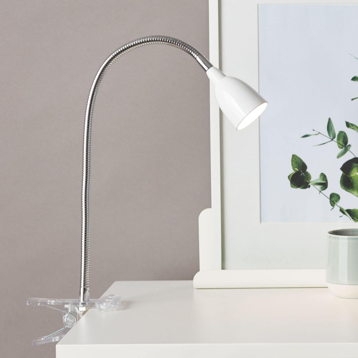 Lampe à pince fixée sur un bureau blanc, éclairant une plante et un cadre décoratif.