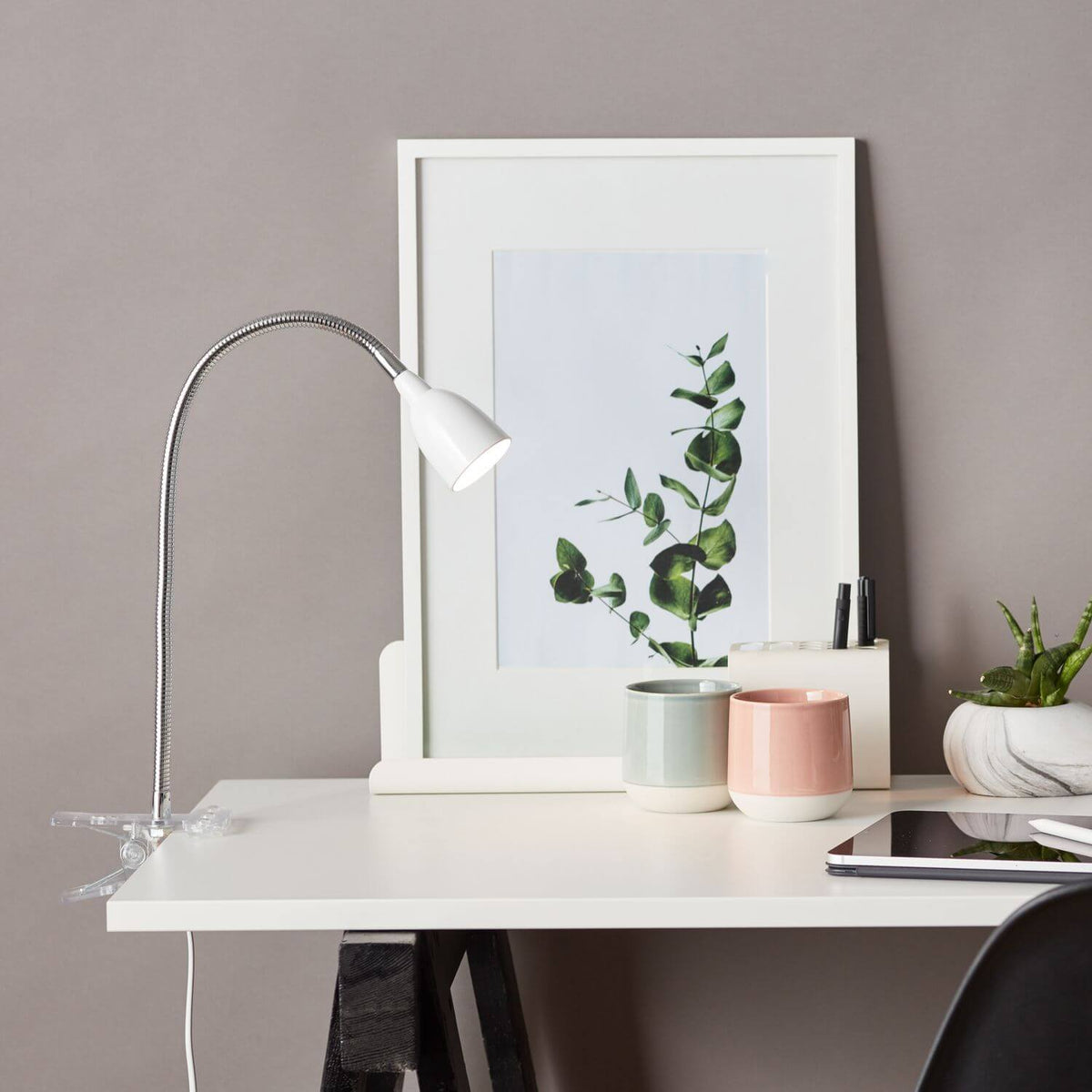 Lampe de bureau à pince blanche éclairant un espace de travail avec cadre, plante et tablette.
