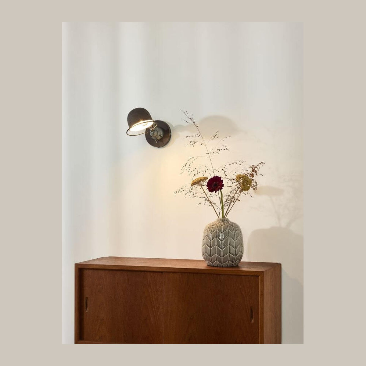 Applique murale allumée au-dessus d'un meuble en bois avec vase de fleurs séchées.