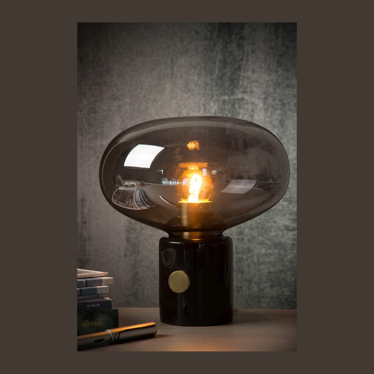 Vue de face du luminaire allumé, socle noir brillant, fond gris texturé.