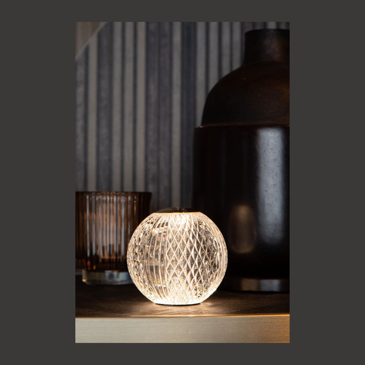 Lampe à poser sphérique en verre transparent facetté allumée, capuchon doré, posée sur une table.