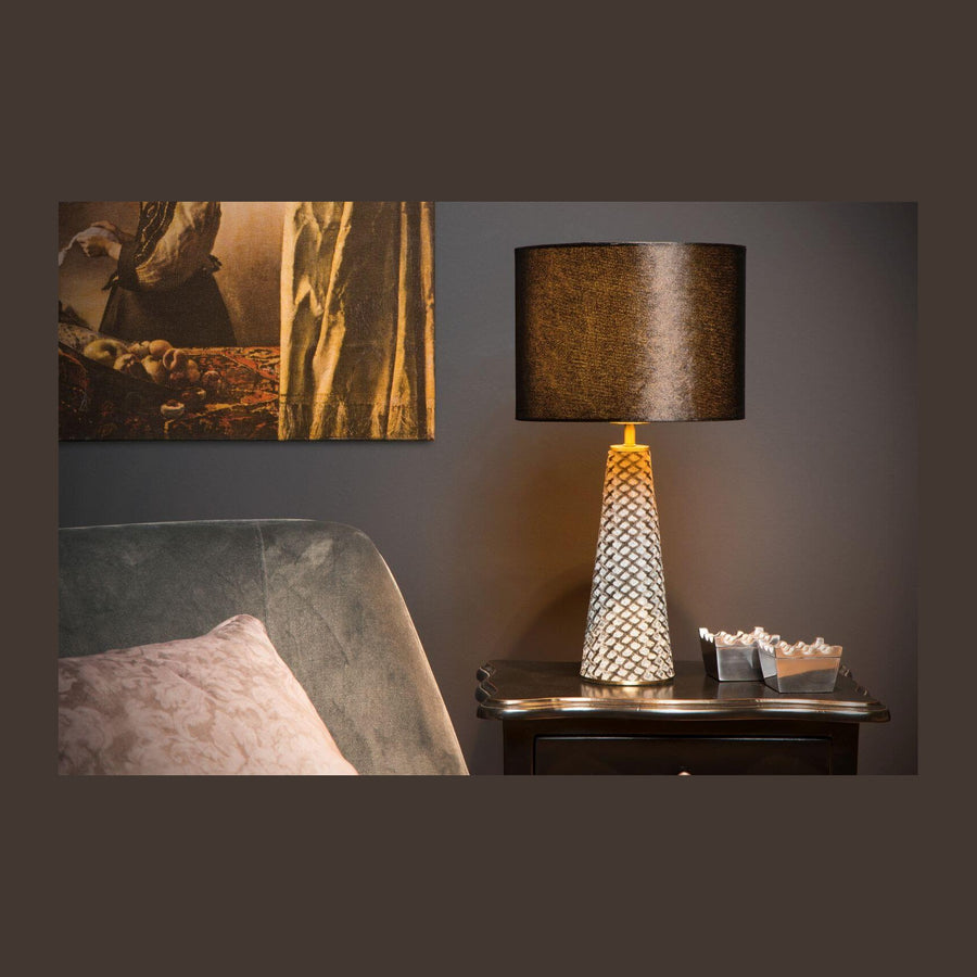 Lampe à poser allumée sur une table de chevet, intérieur sombre avec tableau et lit.
