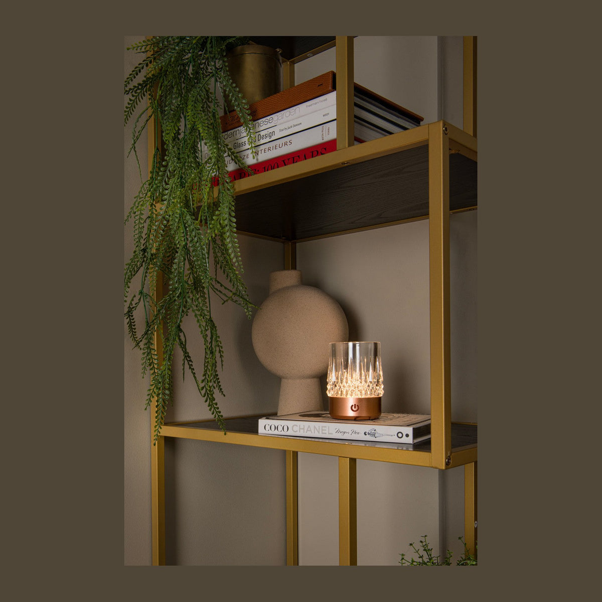 Lampe à poser cylindrique cuivrée allumée sur une étagère, entourée de livres et d'une plante.