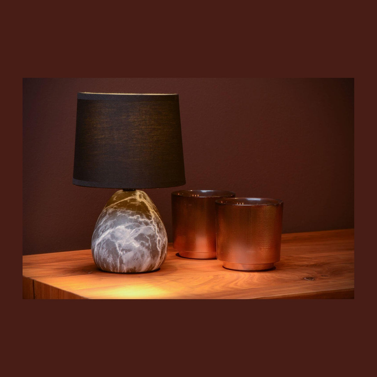 Lampe à poser noire et blanche posée sur une table en bois, avec deux photophores cuivrés.