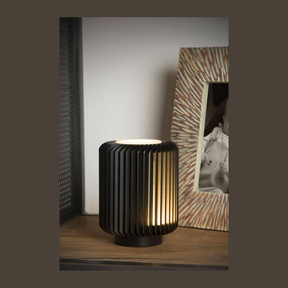 Lampe à poser noire allumée sur une étagère en bois, près d'un cadre photo.