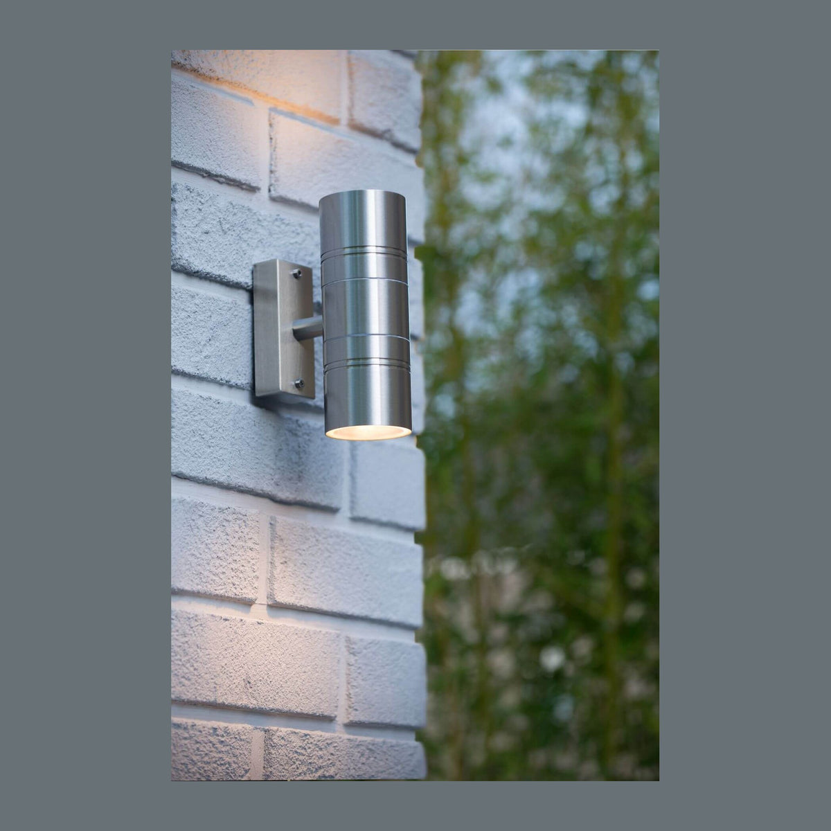 Vue de face d'une applique murale cylindrique en inox sur un mur extérieur.