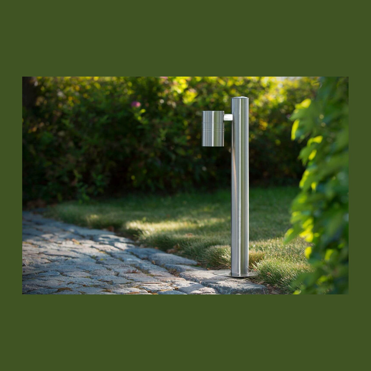 Borne lumineuse extérieure en inox sur une allée en pierre, entourée de verdure.
