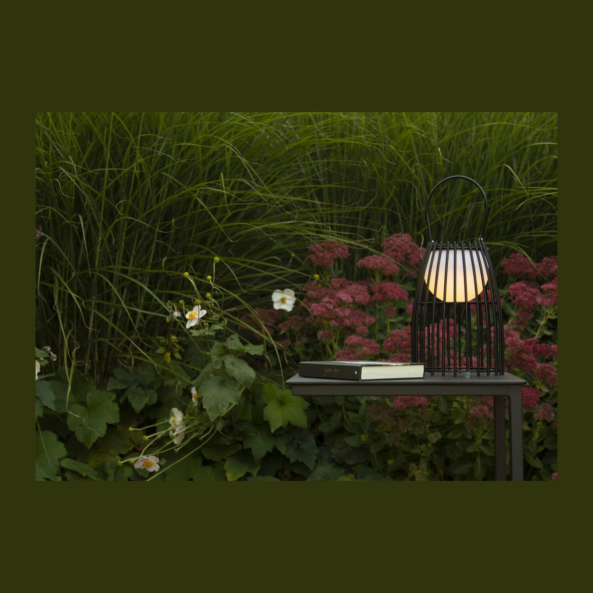 Lampe allumée posée sur une table basse dans un jardin, près d'un livre et de graminées.