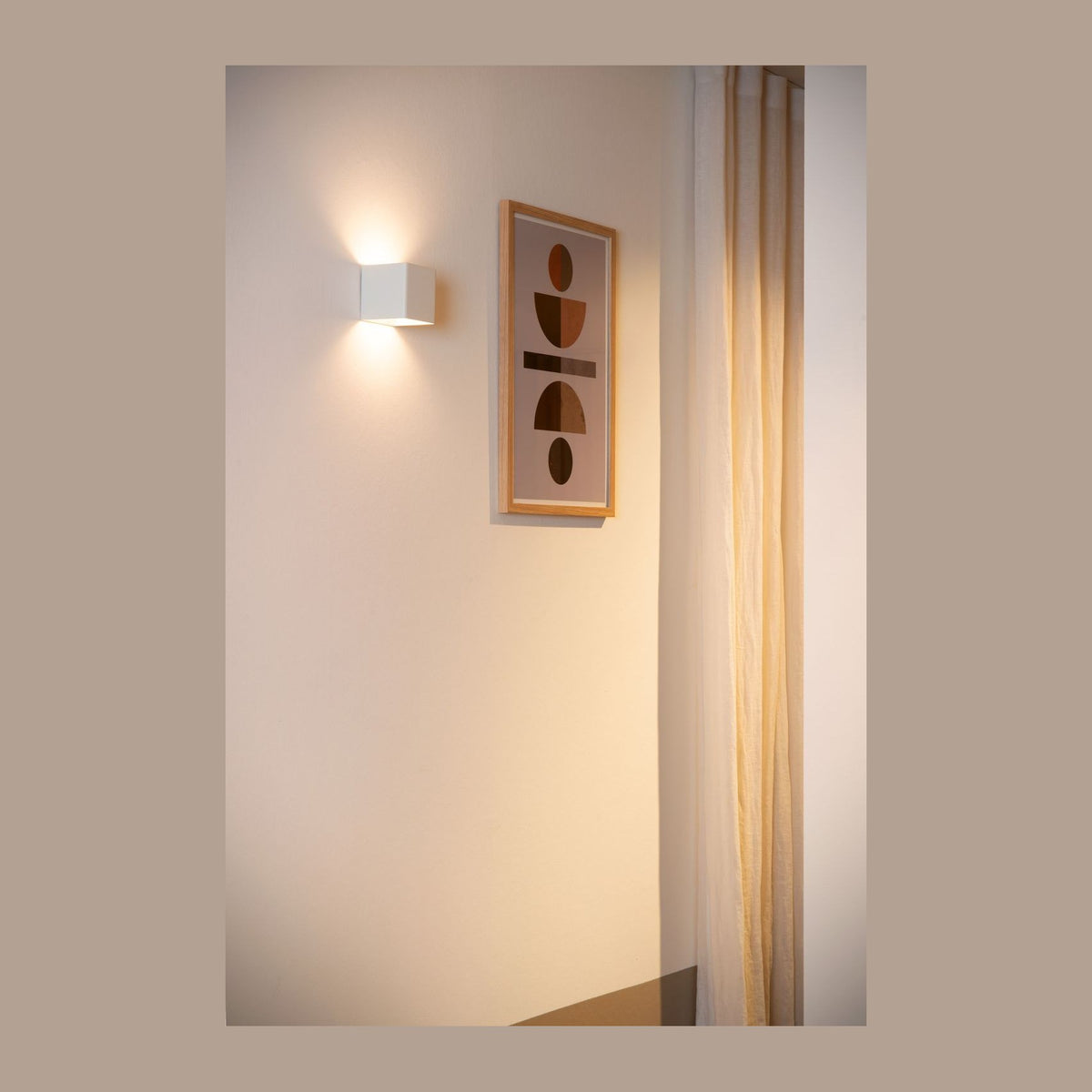 Applique murale blanche allumée sur un mur blanc, près d'un cadre et d'un rideau beige.