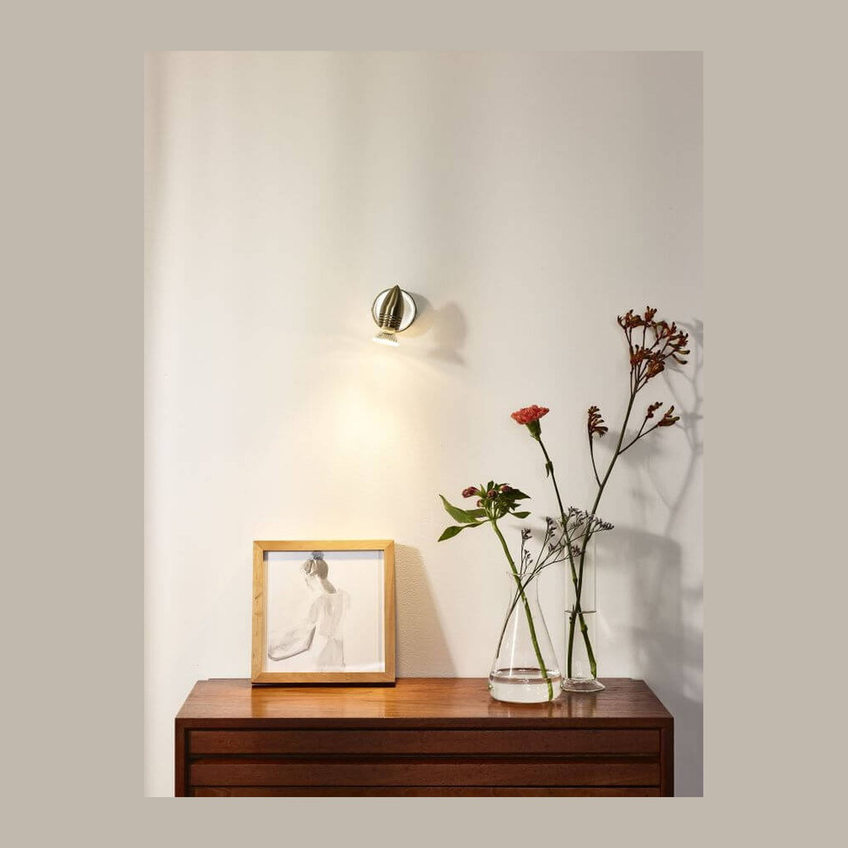 Spot chromé fixé au mur éclairant une commode en bois avec cadre et vase.