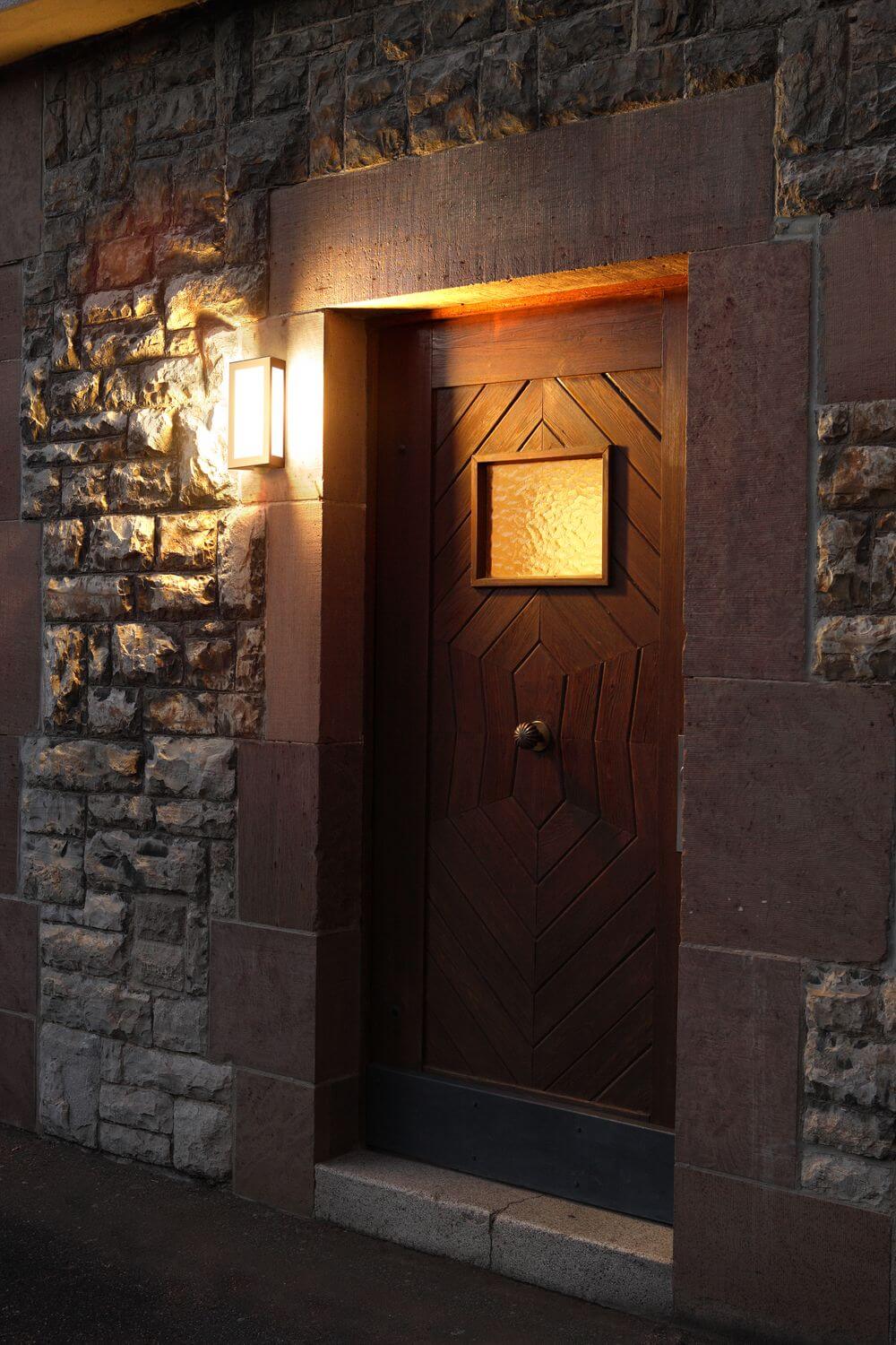 Mise en ambiance extérieure, applique allumée éclairant une porte en bois.