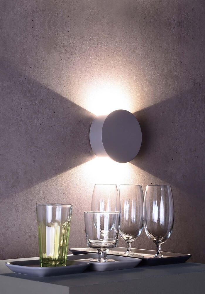 Mise en ambiance de l’applique ronde éclairée, avec verres devant sur un meuble.