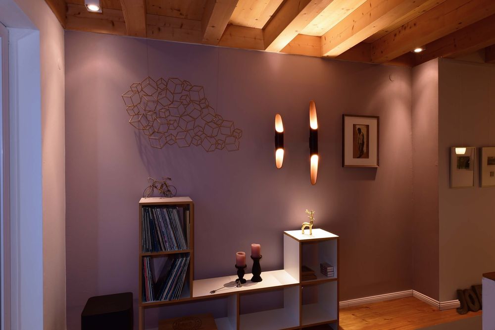 Mise en ambiance sur mur mauve avec deux appliques verticales allumées près d’étagères.